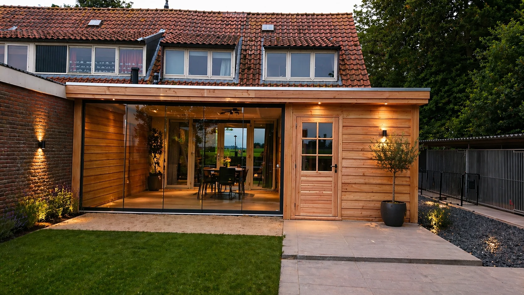 Houten aanbouw met glazen schuifwand tegen een woning, warm verlicht bij schemer