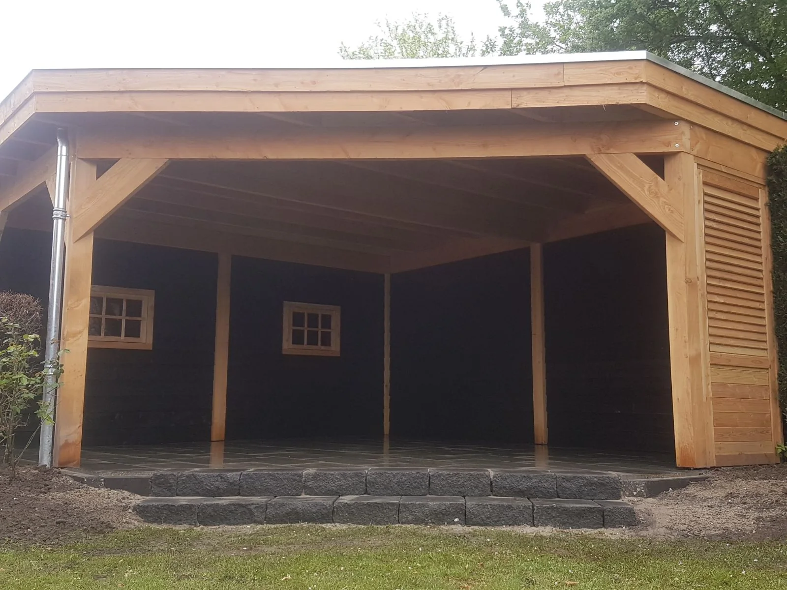 De ruime overkapping met zwarte achterwand en houten raampjes met roeden, op een natuurstenen vloer